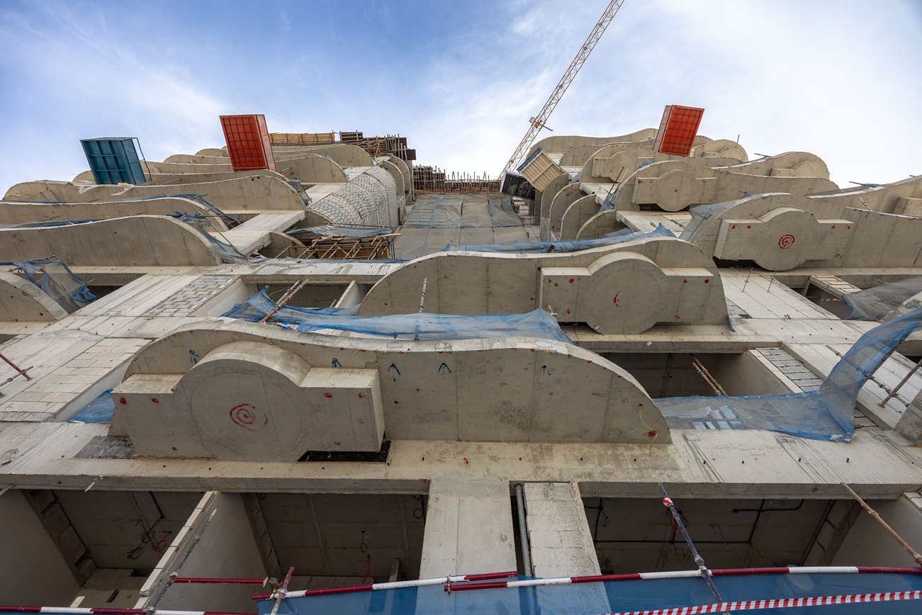 Beach Walk Residence Construction site view showing a building framework with concrete shapes, safety nets, and a crane in the background.